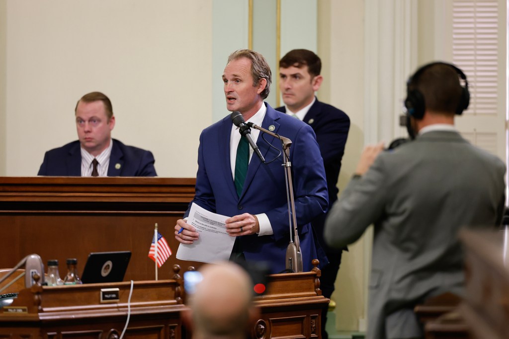 A lawmaker in a blue suit speaks into a microphone while holding papers during a floor session at the California Assembly. Other legislators and staff sit and stand nearby as a photographer records the moment.