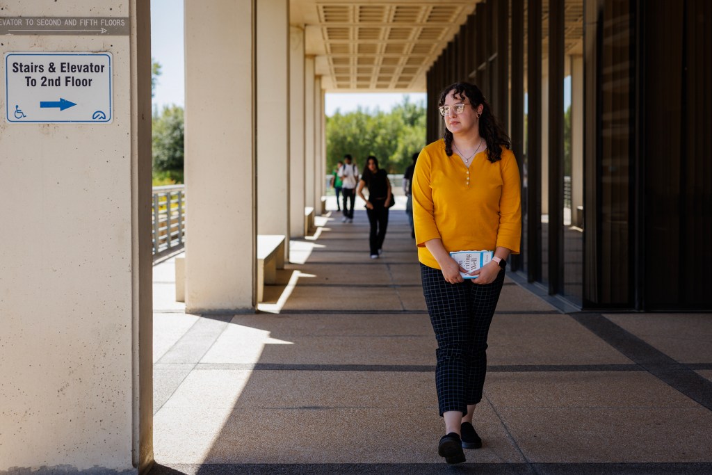 A person in a yellow long-sleeve shirt and checkered pants walks down a shaded outdoor walkway holding a book, with other people visible in the background.